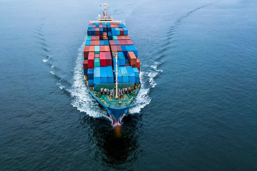 USCanada Tariff Escalation Reshapes North American Supply Chains as Global Freight Costs Surge SYZ Rod Ends Top-down view of a cargo ship at sea, with colorful shipping containers stacked on deck and a visible wake behind it.